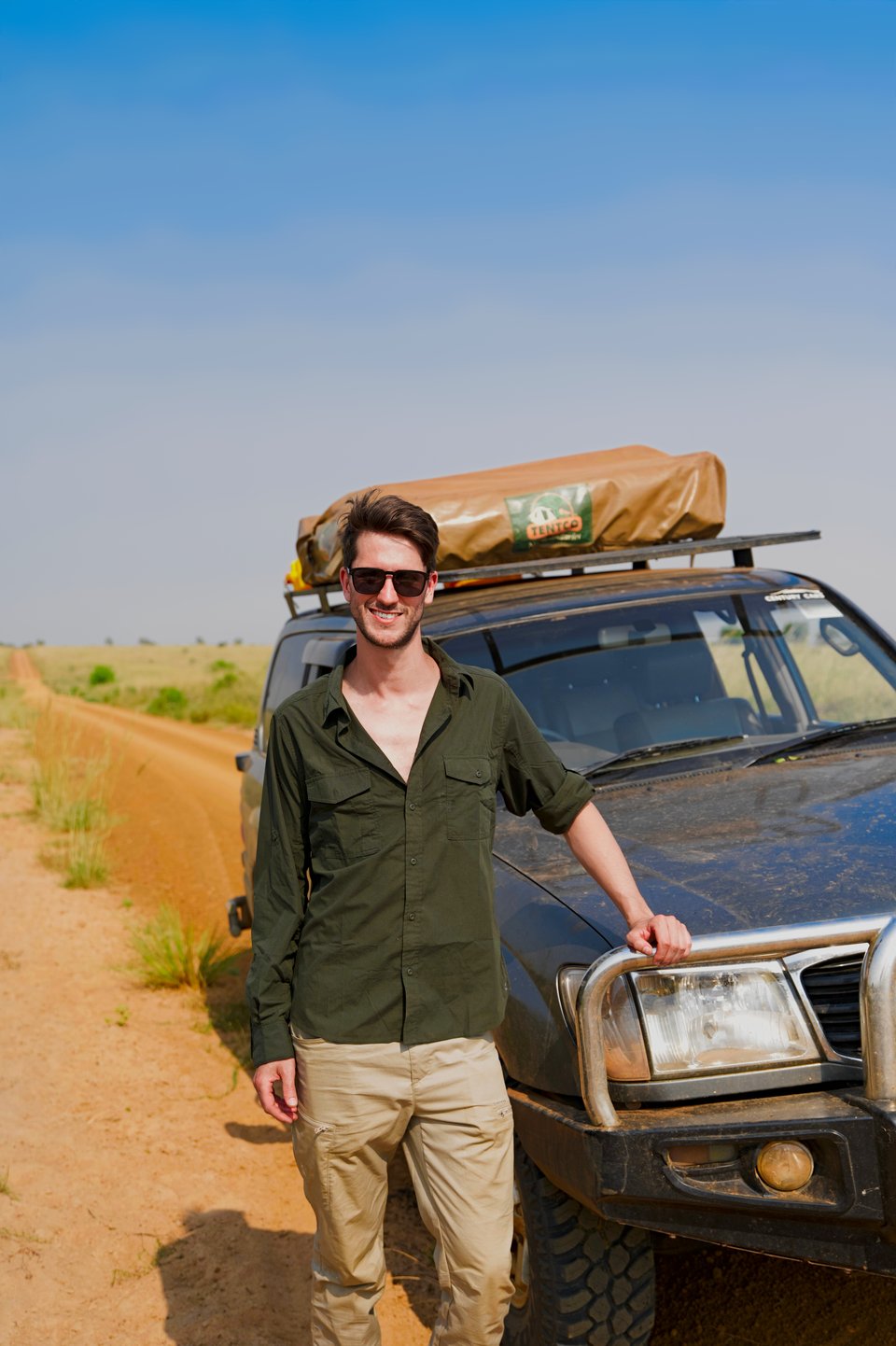 Martijn opiera się o orurowanie 4x4 z namiotem dachowym, obok czerwonej gruntowej drogi przez łąki Ugandy.